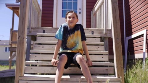 8f9a9c43f7773b9623d1b501f0566a943835ff7b A young person with dark hair sits on wooden steps of a red house, wearing a tie-dye shirt and shorts.
