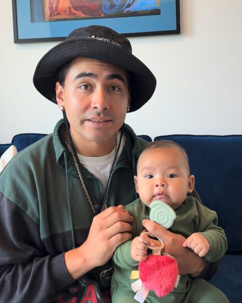 Man Wearing Bucket Hat Green Jacket Holds Baby A man wearing a bucket hat and green jacket holds a baby wearing a green onesie and holding a red apple toy and a green pacifier.