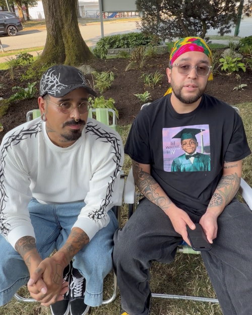 Two Men Sit Outside Chairs Man Left Wears Two men sit outside in chairs. The man on the left wears a white shirt and a black cap. The man on the right wears a black t-shirt and a bandana.