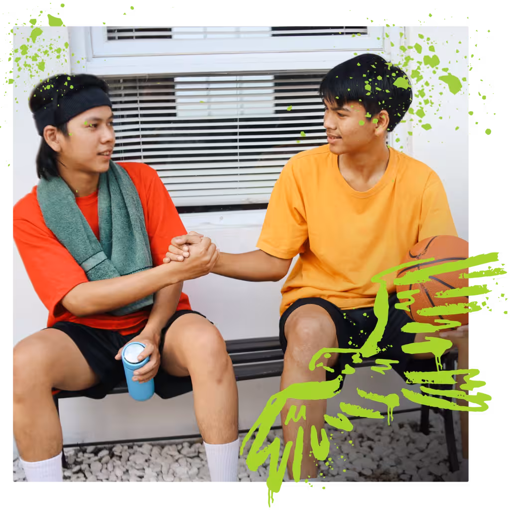 Two young men in athletic wear are sitting on a bench, shaking hands, with one holding a basketball.
